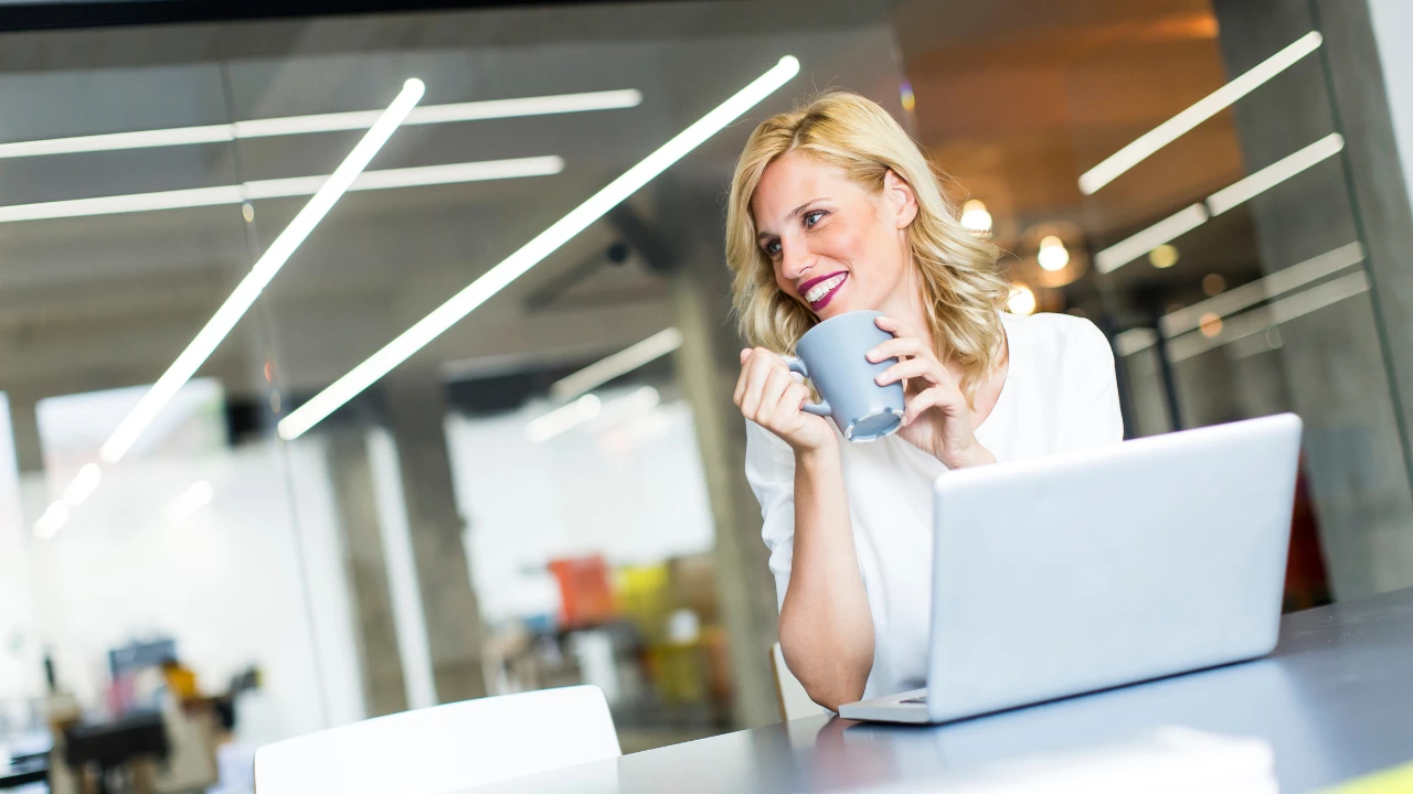 Une femme qui boit son café devant son ordinateur dans un openspace. Elle prend sa pause au travail. 