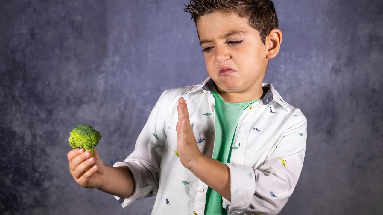 Garçon face à un brocolis qu'il tient dans sa main droite et qui ressent du dégoût en regardant le légume